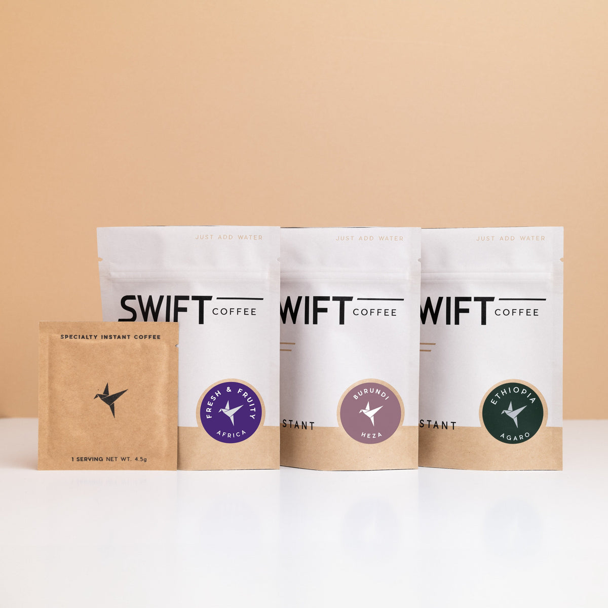Swift Coffee East Africa Trio: Fresh & Fruity Africa, Burundi Heza and Ethiopia Agaro instant coffee pouches with a kraft single-serve sachet on a tan backdrop