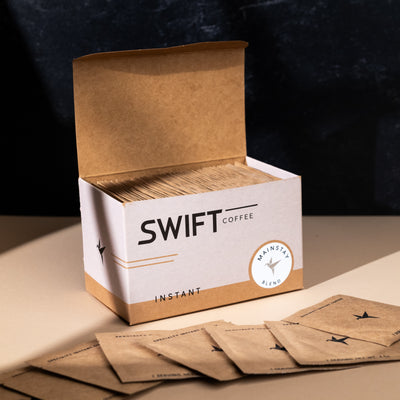 Open box of Swift Coffee Mainstay Blend single-serve sachets with loose sachets spread in front, on a dark backdrop